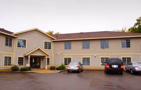 Two-story beige senior living building with a central entrance and parked cars in front.