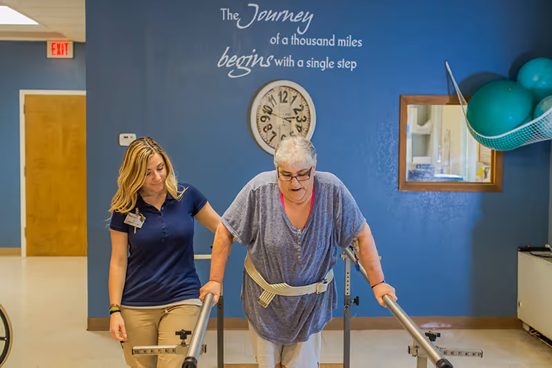 An elderly woman using parallel bars for physical therapy, assisted by a female healthcare worker in a rehabilitation room with a blue wall featuring a clock and an inspirational quote that reads 'The journey of a thousand miles begins with a single step.'