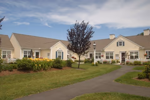 A well-maintained outdoor area of a senior living facility with beige single-story buildings featuring white trim and blue shutters. There is a paved walkway curving through green lawns, flower beds with yellow and orange flowers, and a small tree in the center. The sky is partly cloudy with patches of blue.