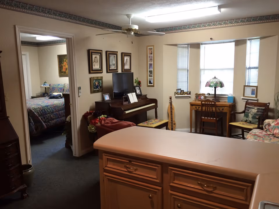 Interior view of a senior living facility room showing a cozy living area with a piano, framed pictures on the wall, a desk with a chair and lamp by the window, and a floral couch. To the left, there is an open doorway leading to a bedroom with a bed covered in a colorful quilt. The foreground shows a kitchen counter with cabinets.