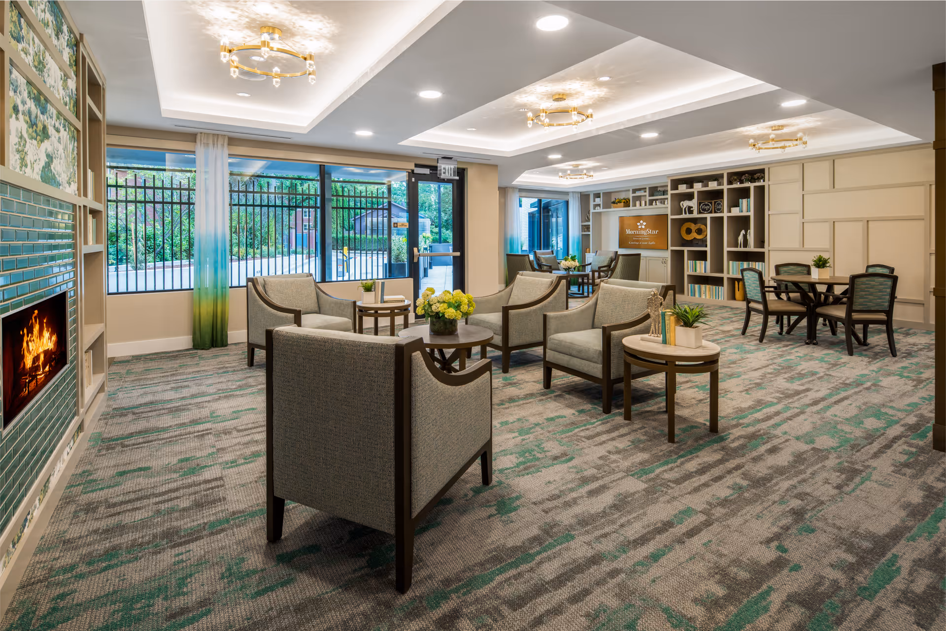 Bright, modern assisted living lounge with upholstered chairs and tables, a fireplace, shelving, and large windows to the outside.