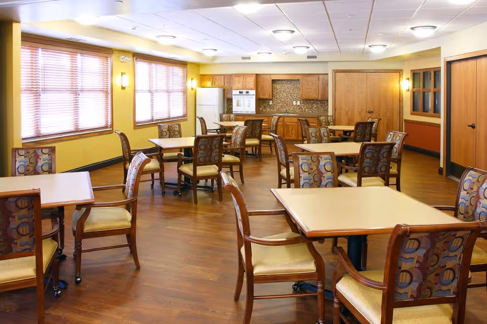 A spacious dining room with multiple square tables and cushioned chairs arranged neatly on a wooden floor. The room has yellow walls, large windows with blinds, and a kitchen area with wooden cabinets, a refrigerator, and an oven in the background.