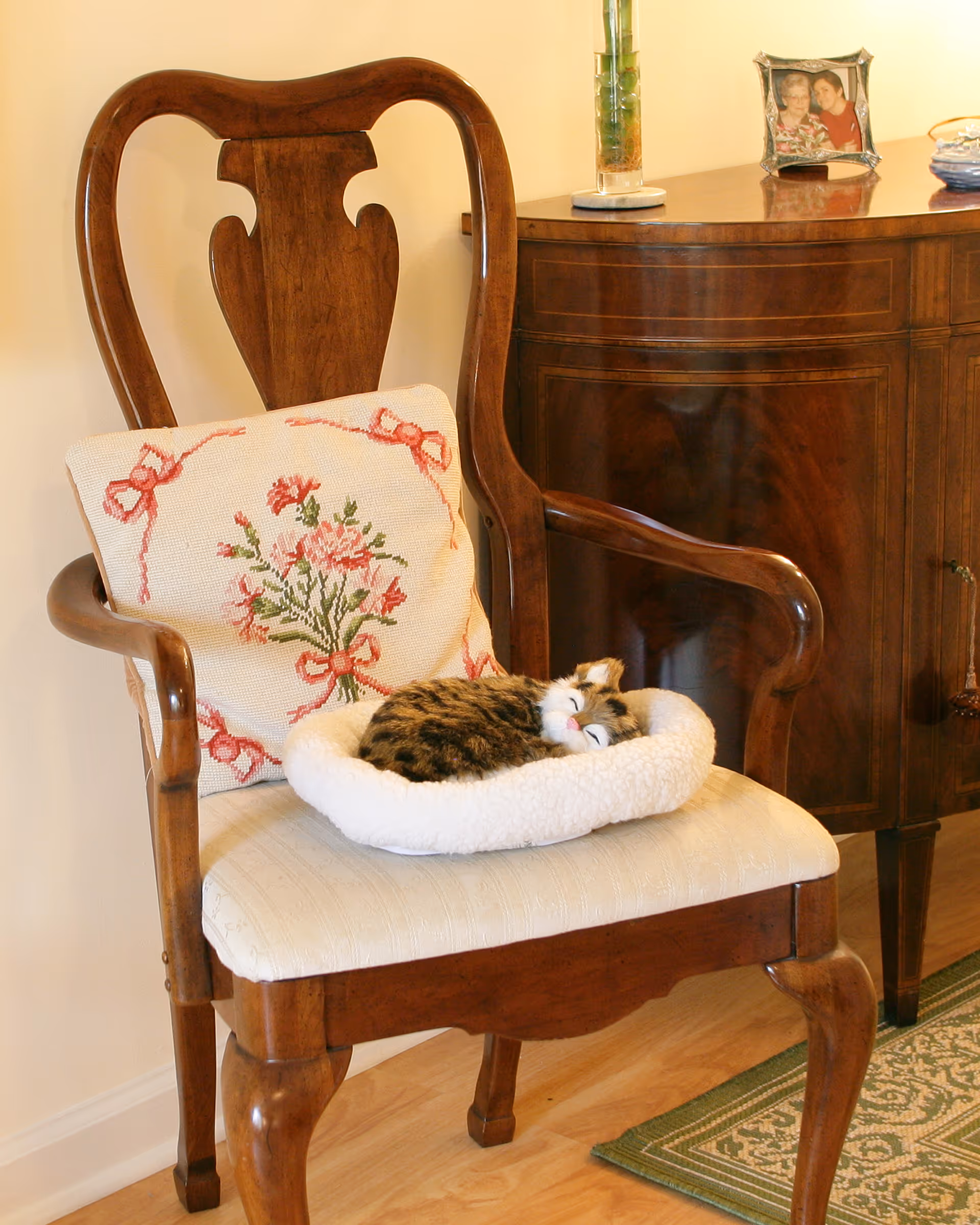 A wooden armchair with a cream-colored cushioned seat and a decorative pillow featuring a floral design. A small tabby cat is curled up and sleeping on a soft white pet bed placed on the chair. In the background, there is a wooden cabinet with a framed photo and a vase on top.