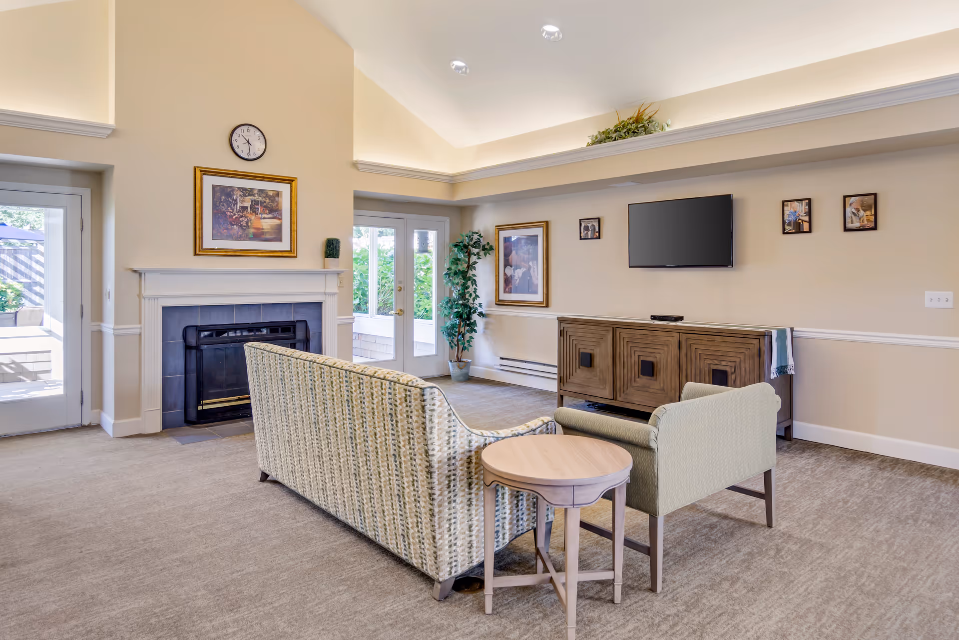 Bright communal living room with a sofa and armchair facing a wall-mounted TV, fireplace, and glass doors to the outside.