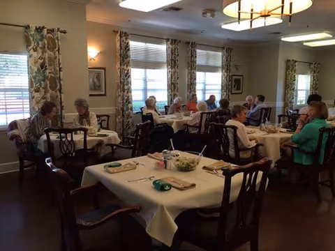 A dining room in Argent Court Assisted Living with several elderly people seated at tables covered with white tablecloths, eating and conversing. The room has floral curtains, framed pictures on the walls, and warm lighting from ceiling fixtures and wall sconces.
