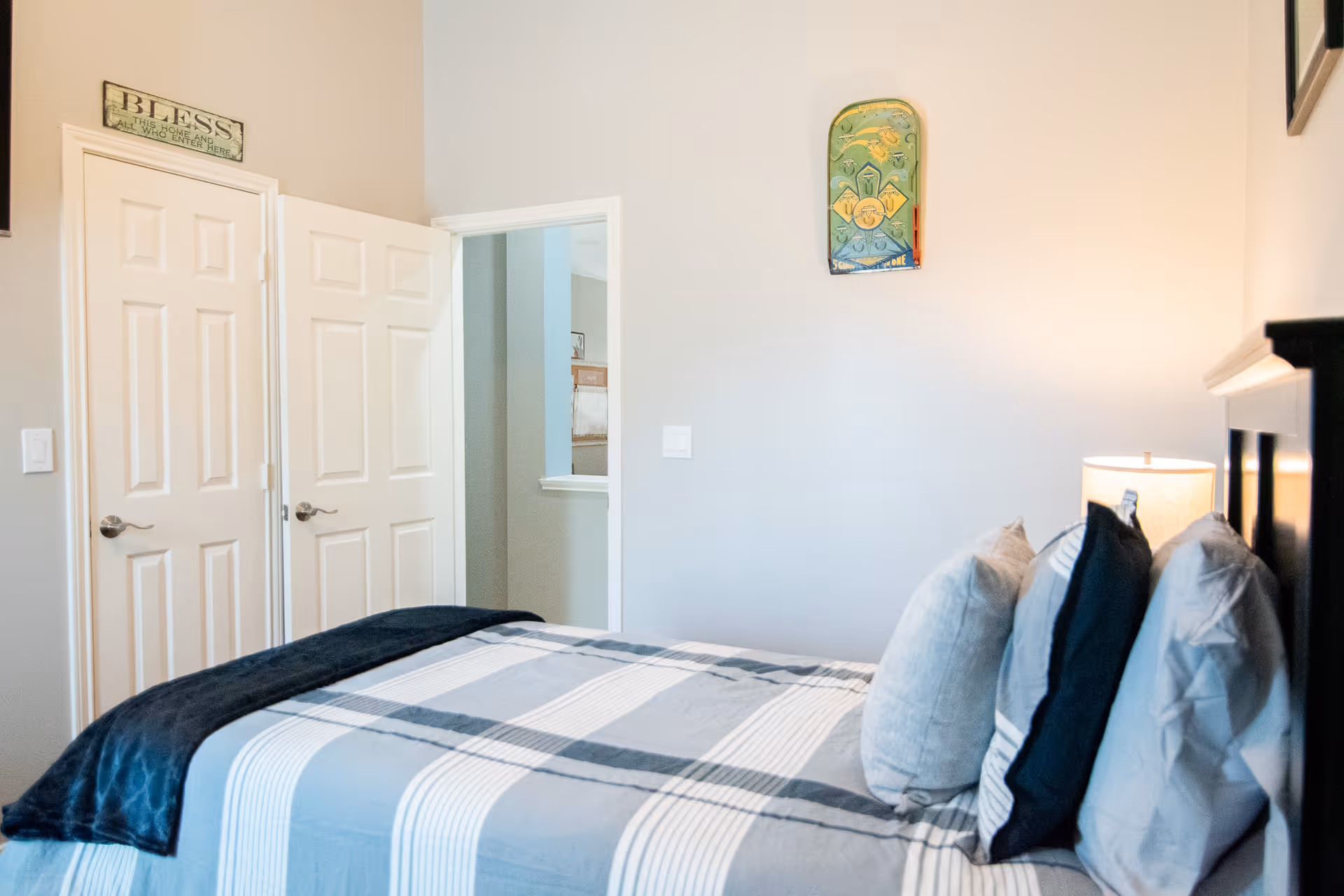 A bedroom with a neatly made bed featuring striped bedding and multiple pillows. There is a lamp on a nightstand beside the bed. Two white doors are visible, one slightly open, and light-colored walls with minimal decorations including a small wall hanging and a sign above one door.