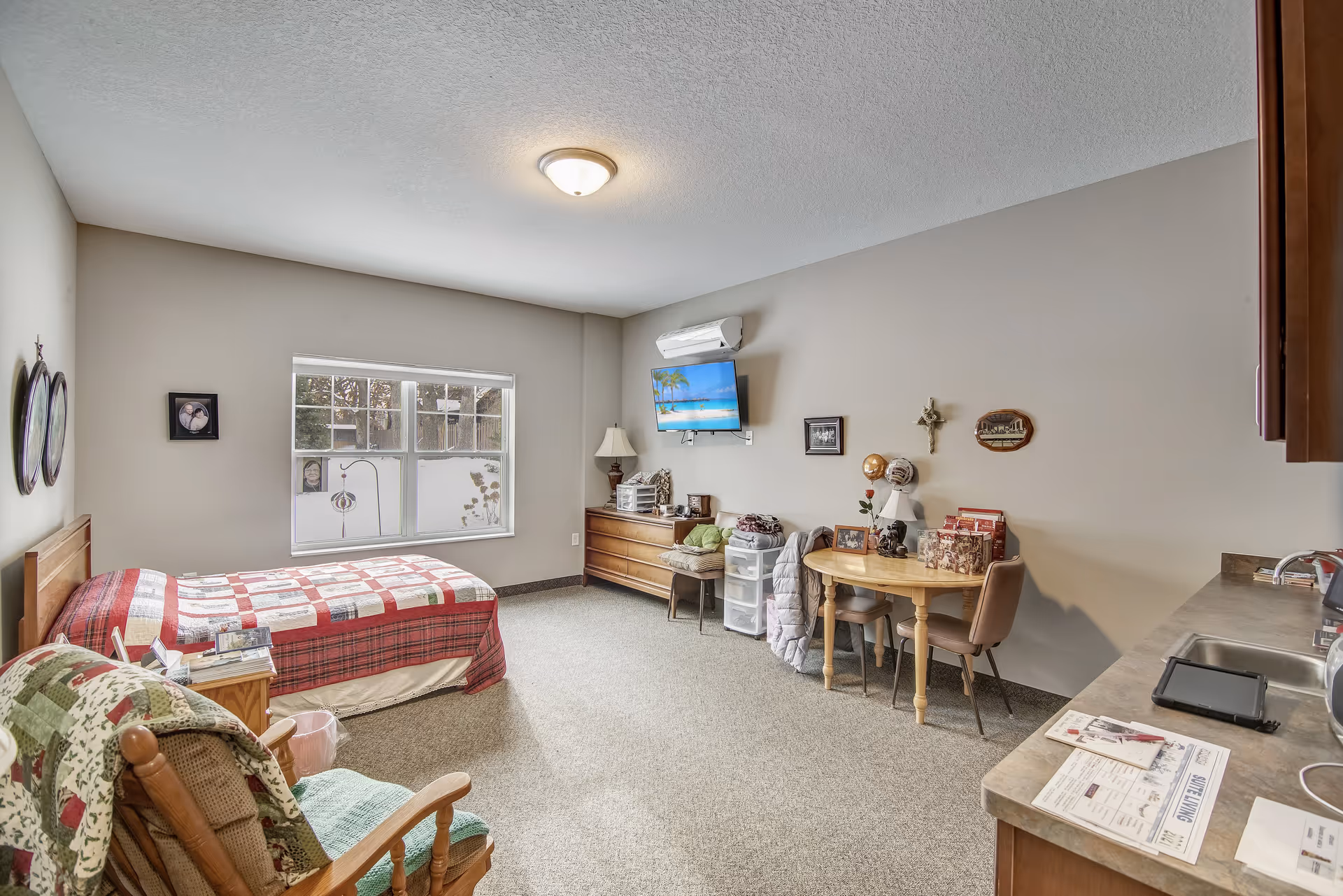 A bright assisted-living bedroom with a twin bed, dresser and wall-mounted TV, small dining table and chairs, kitchenette sink, and a rocking chair.