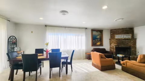 Bright combined living and dining room with a table and chairs, upholstered seating, and a brick fireplace displaying a 'Home of Purpose' sign.
