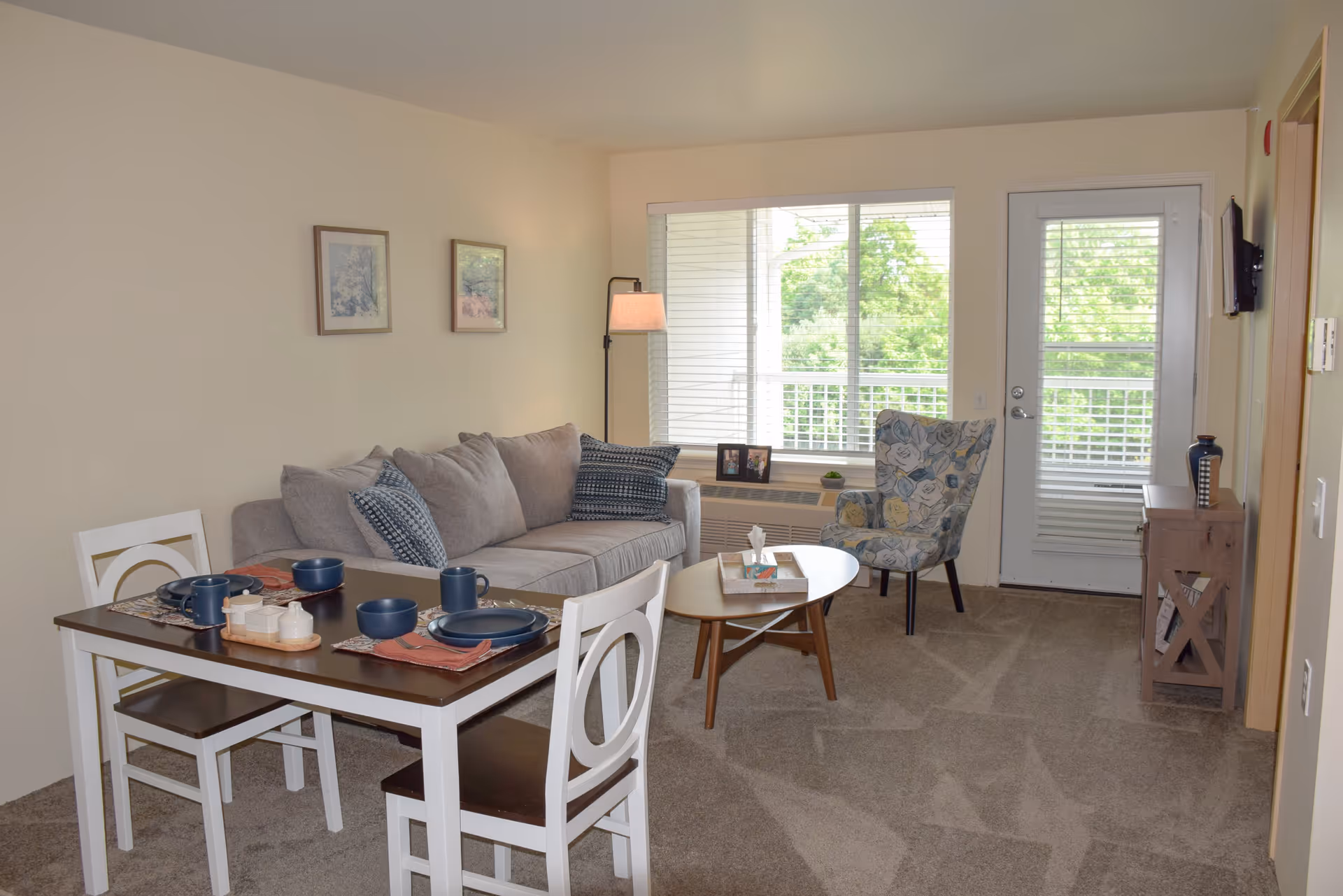 A cozy living room and dining area with a gray sofa adorned with patterned pillows, a floral armchair, a wooden coffee table with books and a tissue box, and a dining table set with blue dishes and placemats. The room has beige walls, carpeted floor, a large window with blinds, a door leading outside, and a floor lamp beside the sofa.