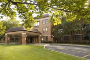 Exterior view of Belhaven Nursing Home, a multi-story brick building with a covered entrance driveway surrounded by green grass and trees with sunlight filtering through the leaves.