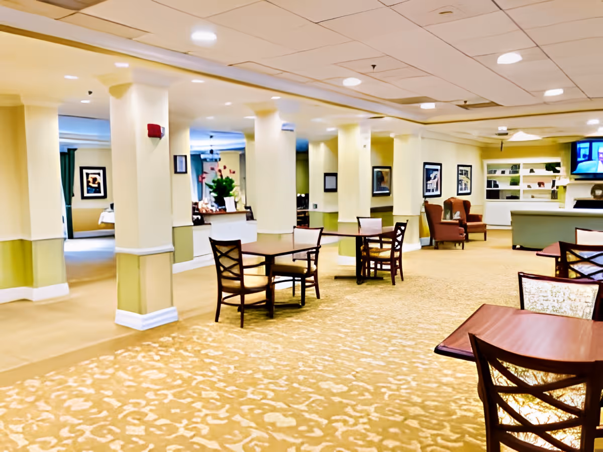 Interior view of a senior living facility common area with several tables and chairs arranged on a patterned carpet. The room has cream-colored walls with green accents and multiple columns. There are framed pictures on the walls, a television mounted in the background, and a reception or service desk area. Comfortable armchairs are placed near the back wall.