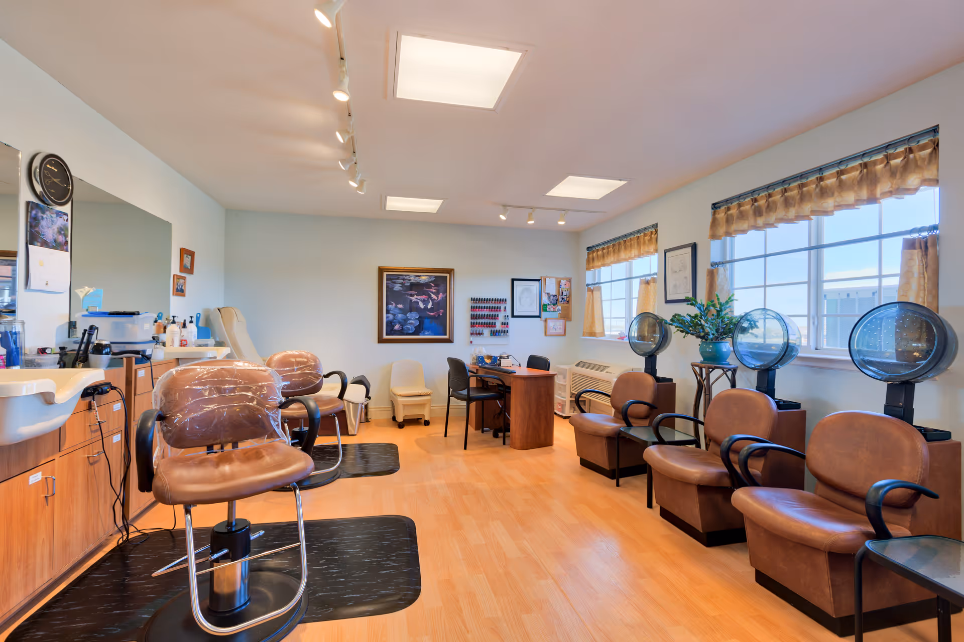 Interior view of a salon area in a senior living facility named Shawnee Hills, featuring salon chairs with plastic covers, hair drying stations, a manicure table with chairs, and large windows with valance curtains letting in natural light.