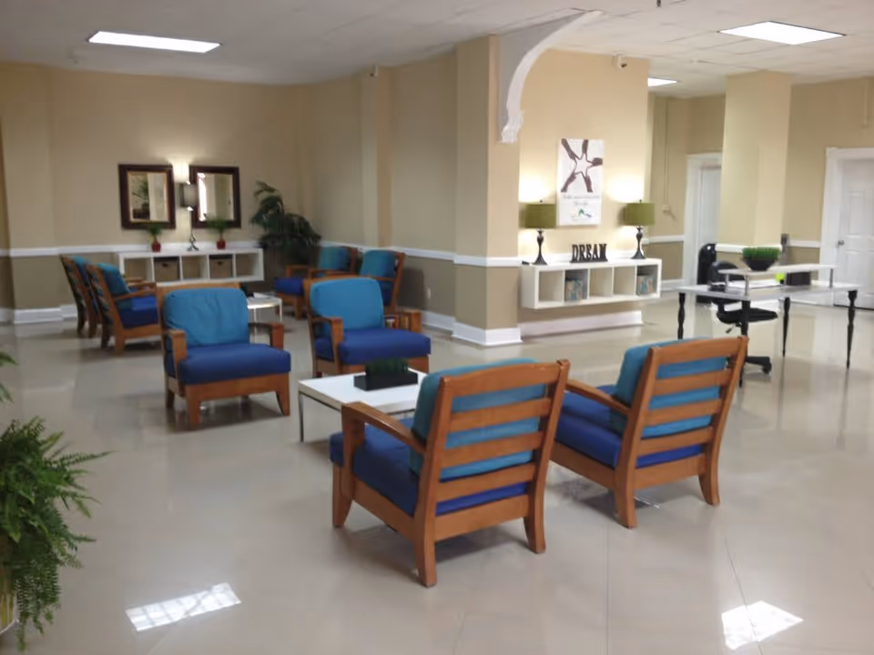 A spacious and well-lit common area with multiple wooden chairs featuring blue cushions arranged around white coffee tables. The room has beige walls with white trim, decorative plants, wall art, and a desk with a black office chair in the background.