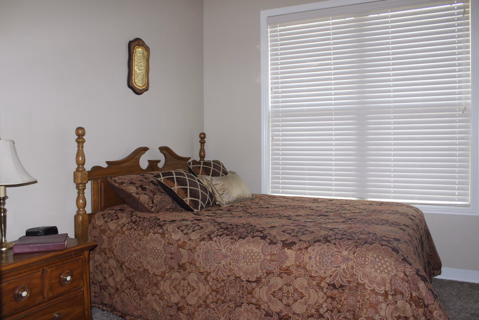 A bedroom with a wooden bed frame and a patterned brown bedspread. There are several decorative pillows on the bed. A wooden nightstand with a lamp and a book is next to the bed. A window with closed white blinds is on the right wall, and a small decorative plaque hangs on the wall above the bed.