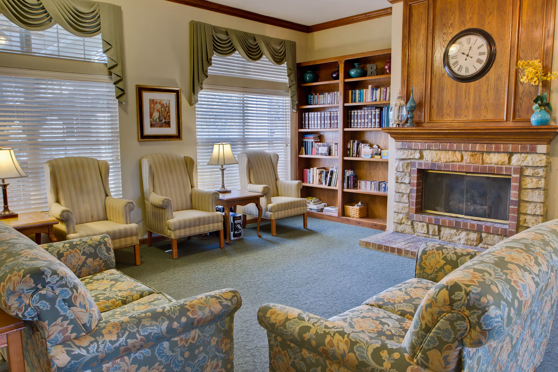 Cozy lounge with floral sofas and armchairs arranged around a fireplace, bookshelf, and lamps.