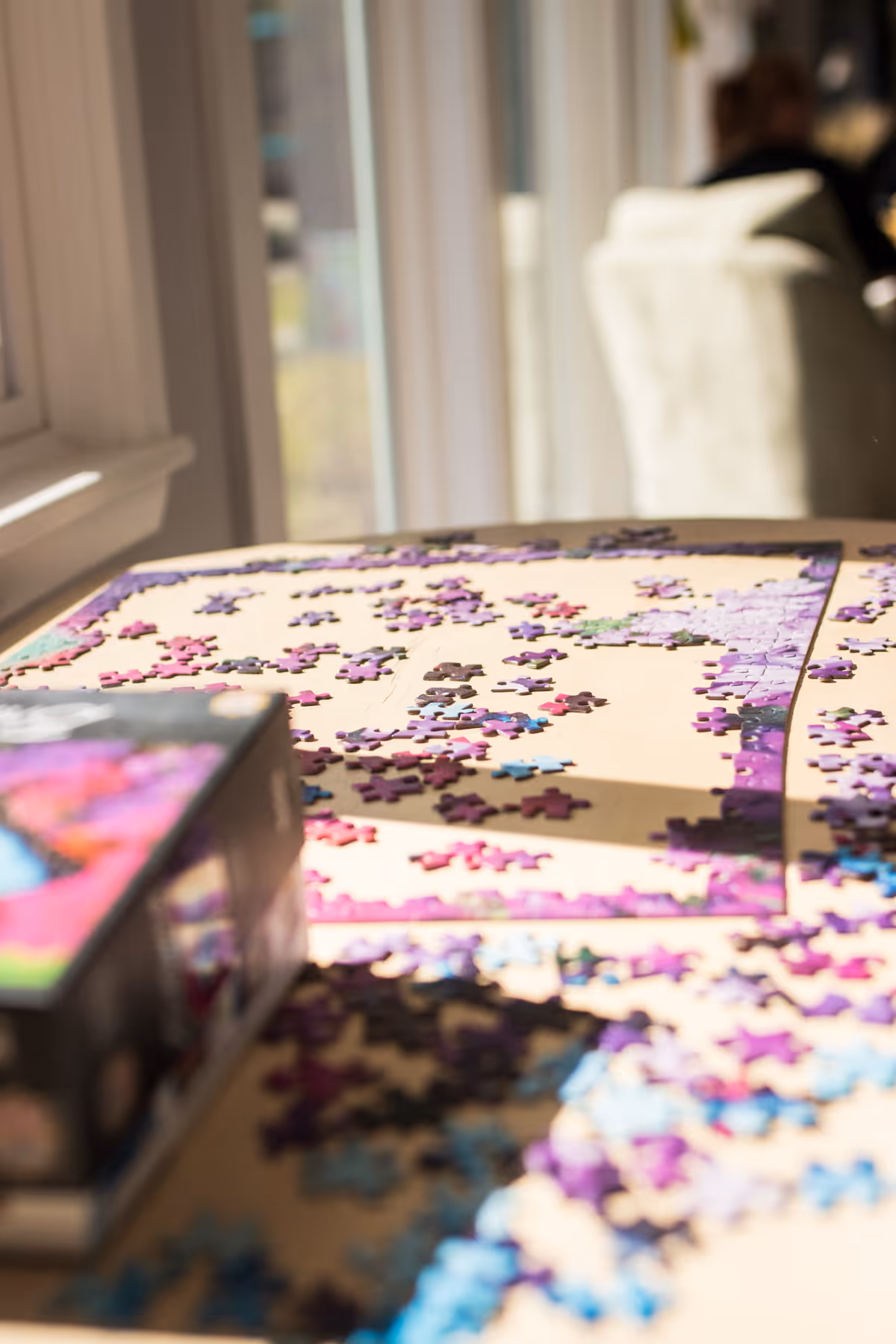 Table by a window covered in scattered colorful jigsaw puzzle pieces and a puzzle box.