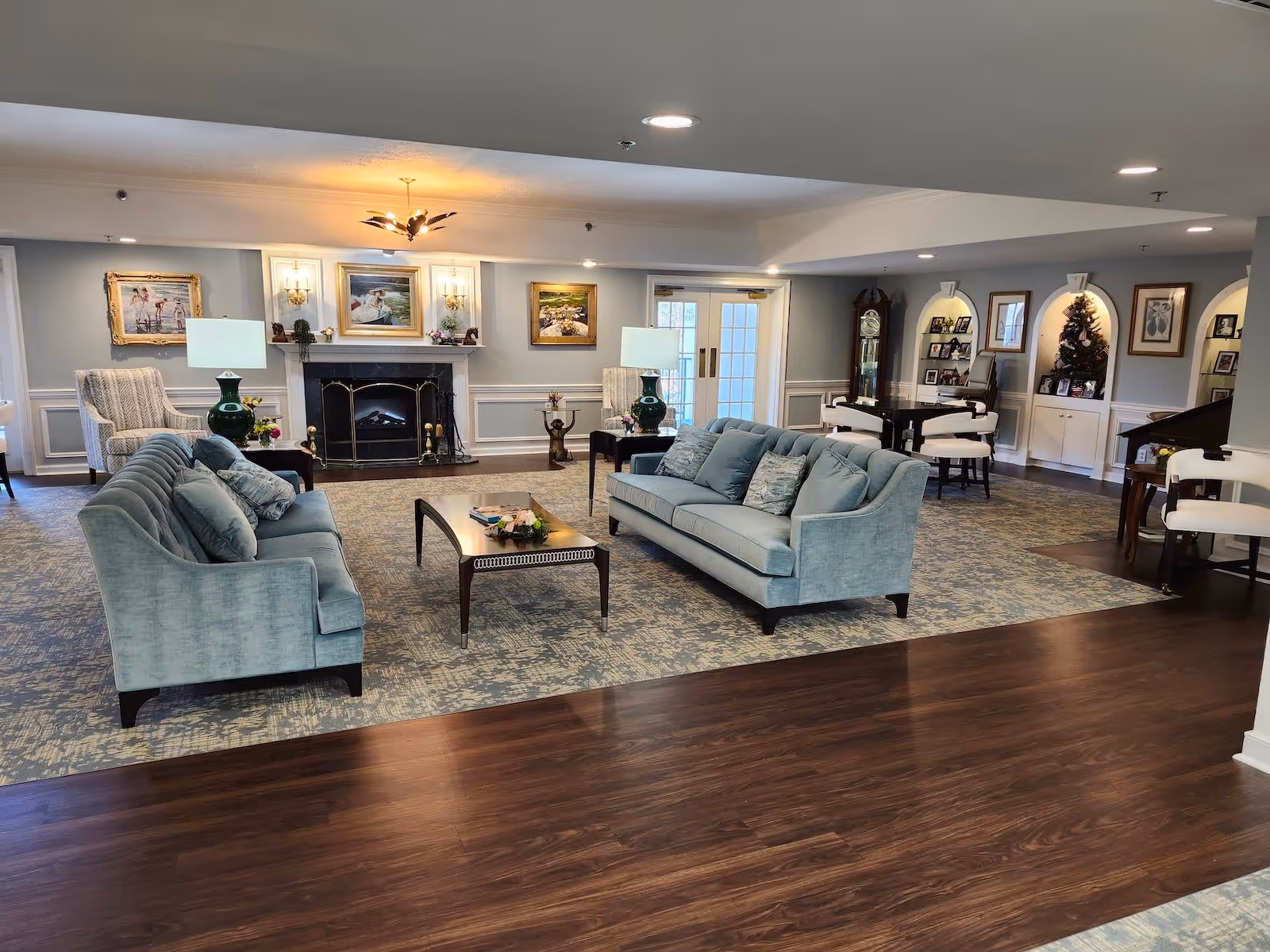 A spacious and elegantly decorated living room area in Cumberland Village featuring two light blue sofas facing each other with a dark wooden coffee table in between. The room has a patterned carpet with wooden flooring around the edges, a fireplace with framed paintings above it, two armchairs with green lamps on side tables, and a corner with a grandfather clock, a small table with chairs, and built-in shelves displaying photos and a small Christmas tree.