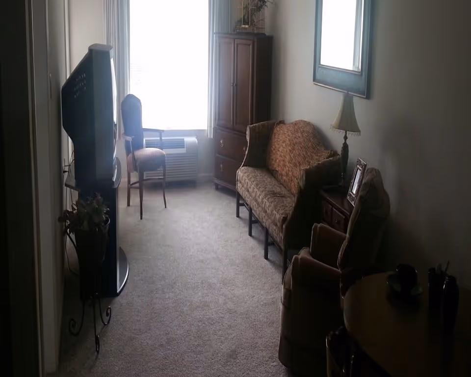 Small living room with a patterned loveseat, armchairs, TV on a stand, wooden cabinet, lamp and a window with an air conditioner.