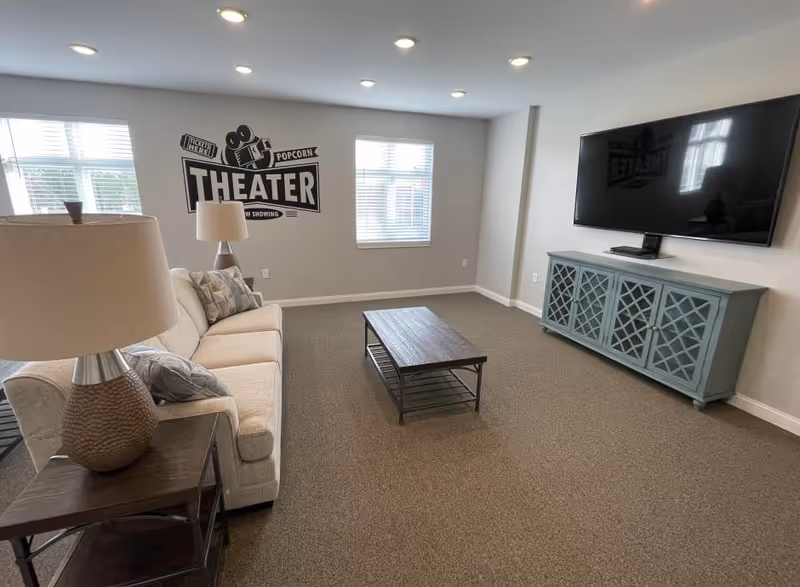 A cozy common living room with a beige sofa, coffee table, large wall-mounted TV, and a 'THEATER' wall decal on the far wall.