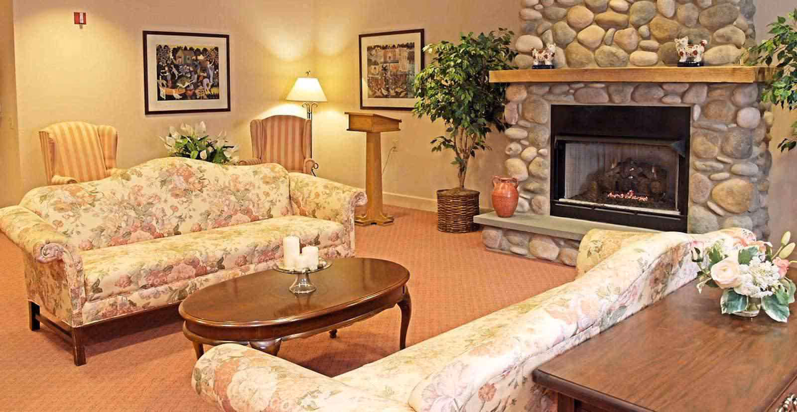 A cozy living room with two floral patterned sofas facing each other, a wooden coffee table with candles, two striped armchairs, a stone fireplace with decorative items on the mantel, framed artwork on the walls, a floor lamp, and potted plants.