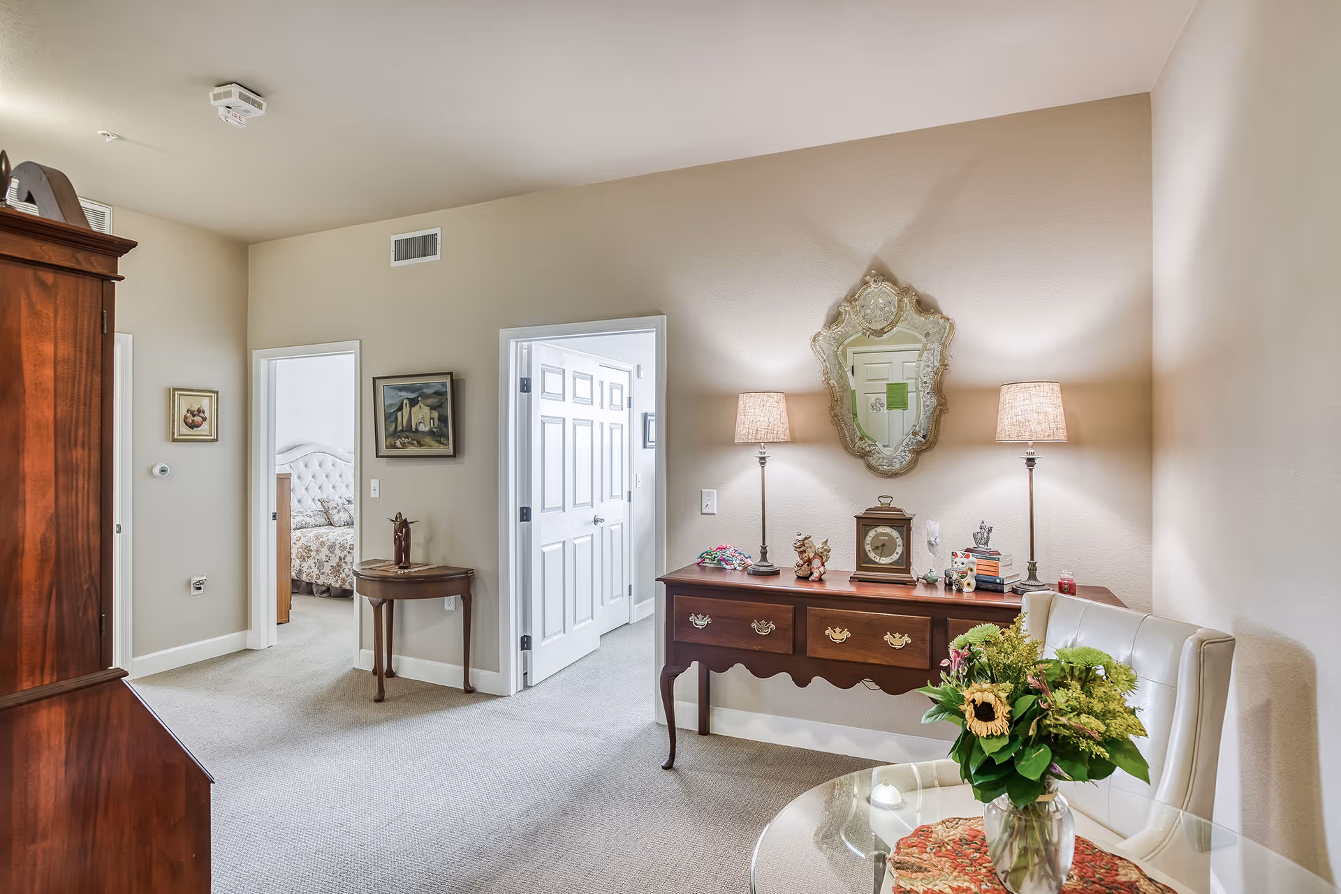 Interior view of a senior living facility room with beige walls and carpet. There is a wooden sideboard with two lamps, a decorative mirror, clock, and small ornaments on top. A glass table with a white cushioned chair and a vase of flowers is in the foreground. Two doorways lead to other rooms, one showing a bed with a tufted headboard. Paintings hang on the walls.