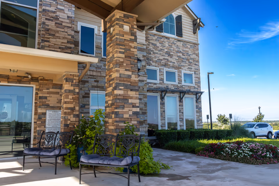 Outdoor seating area at the entrance of a building with stone brick pillars, two black metal benches with cushions, green plants, and a flower bed. A white car is parked nearby under a clear blue sky.