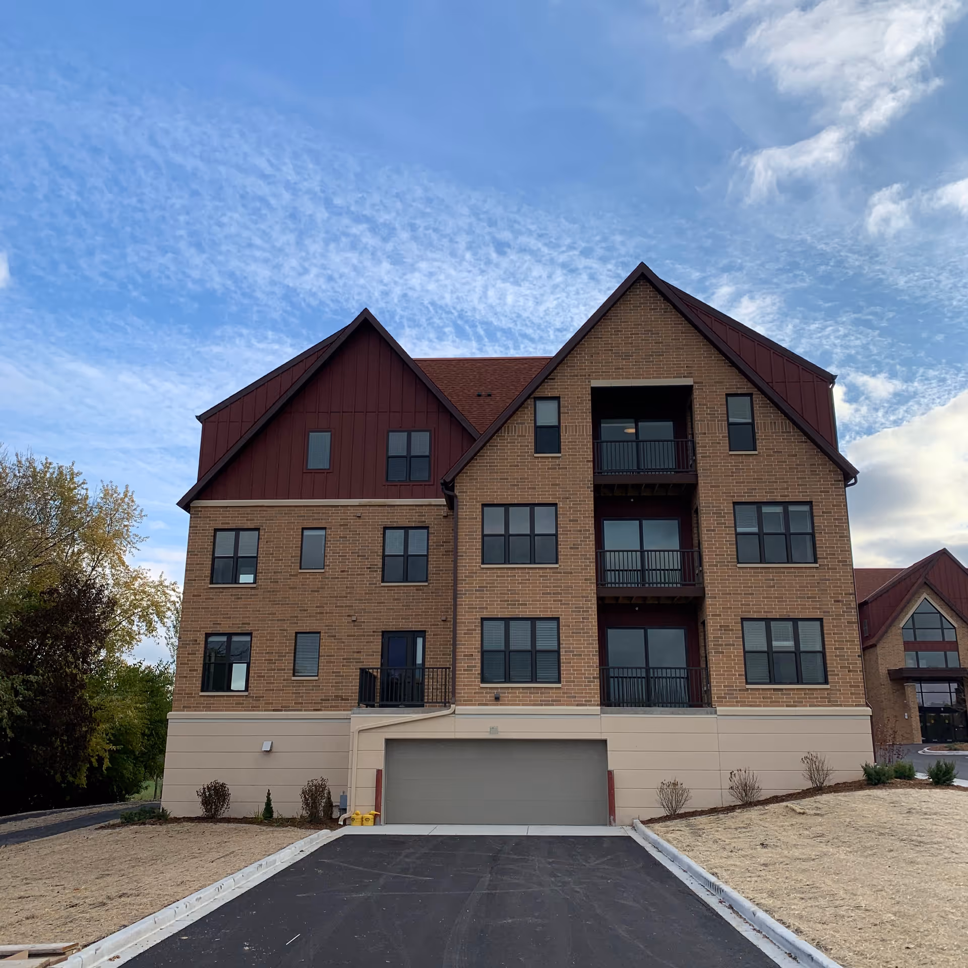 Exterior view of a multi-story brick building with a sloped roof and multiple windows, featuring balconies and a garage entrance at the ground level, under a partly cloudy blue sky.