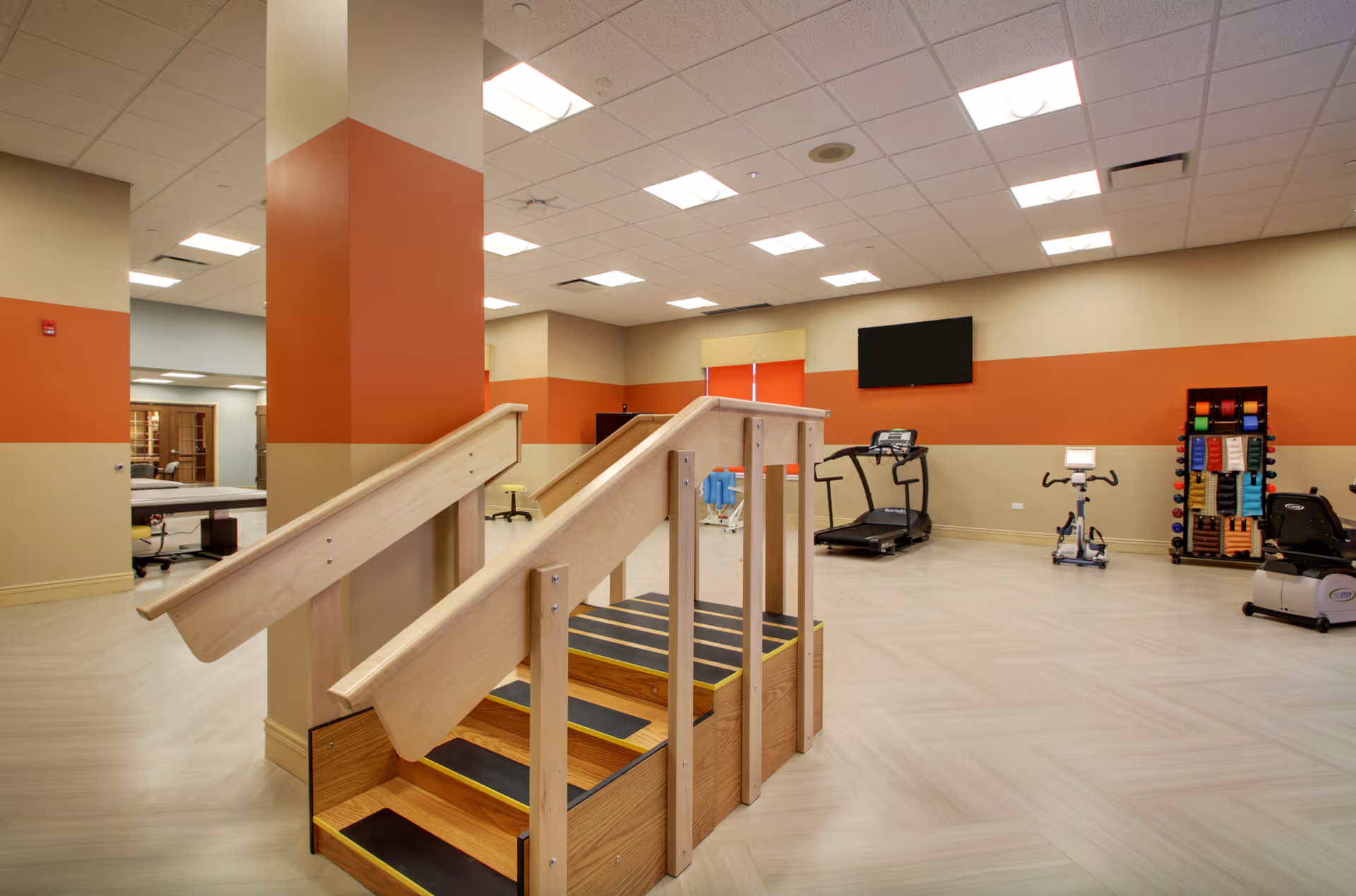A spacious physical therapy or rehabilitation room with a wooden staircase for practice, a treadmill, exercise bike, and a rack of colorful exercise bands and weights. The walls are painted beige with an orange stripe, and there is a large flat screen TV mounted on the wall.
