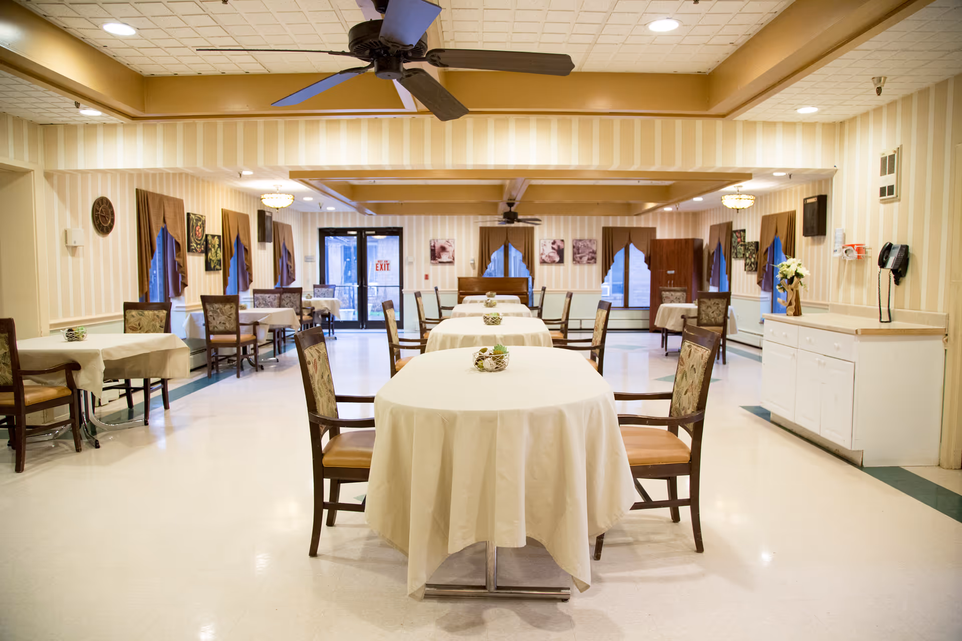 A spacious dining room with multiple tables covered in white tablecloths, each surrounded by wooden chairs with cushioned seats. The room has striped wallpaper, several windows with brown curtains, ceiling fans, and decorative wall art. There is a white cabinet with a telephone and a flower arrangement on the right side of the room.