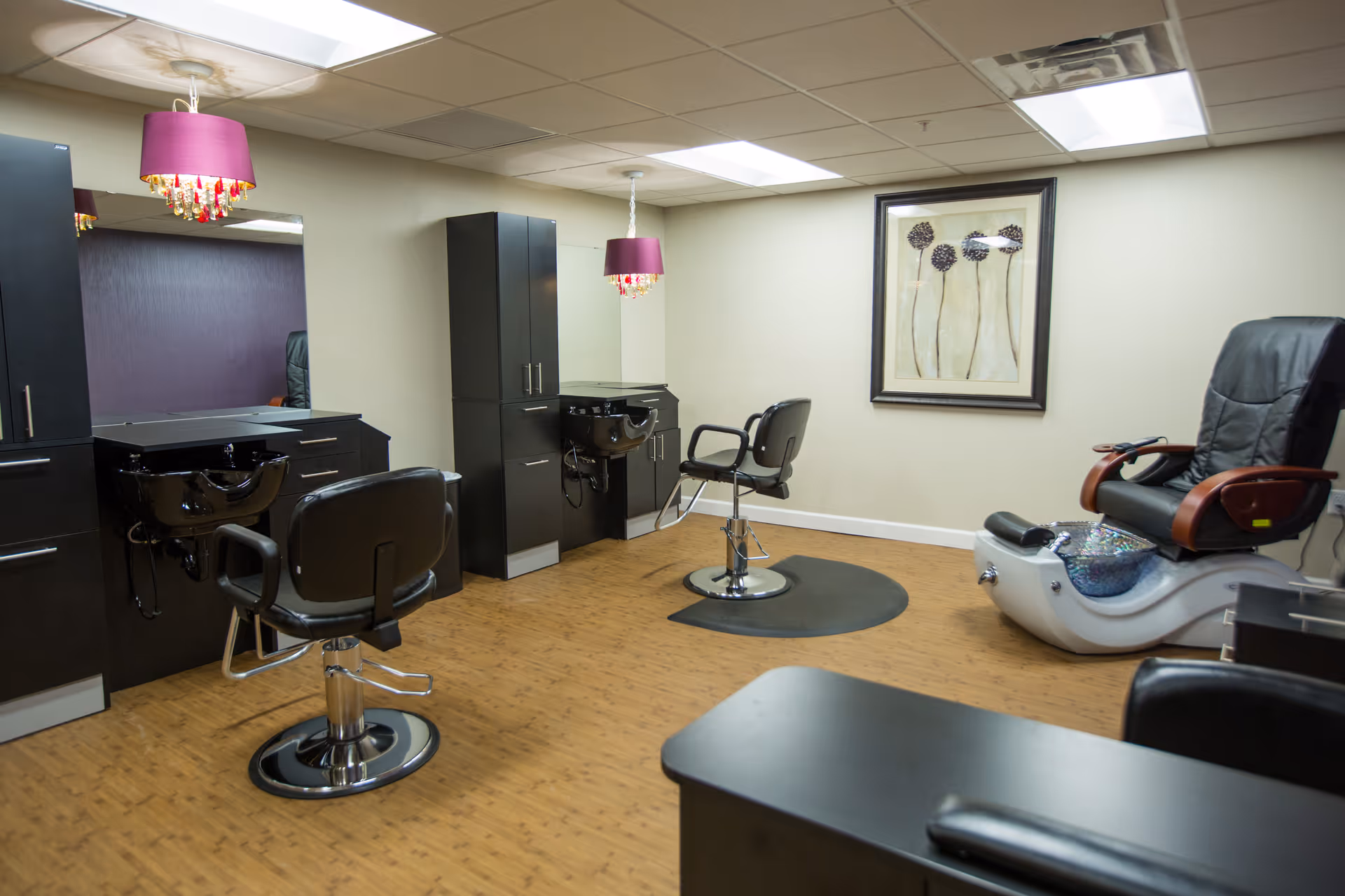 Interior of a salon room with two black salon chairs in front of sinks and mirrors, a pedicure chair with foot bath, wooden flooring, beige walls, a framed floral artwork, and two purple pendant lights hanging from the ceiling.