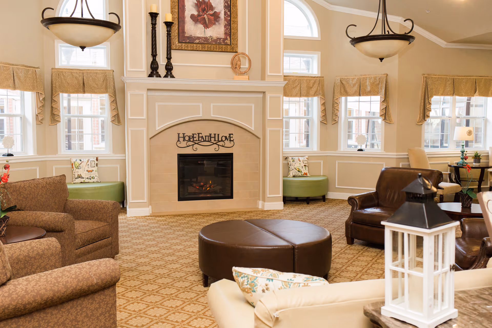 Communal living room with a central fireplace, cushioned seating, a round ottoman, and multiple windows.