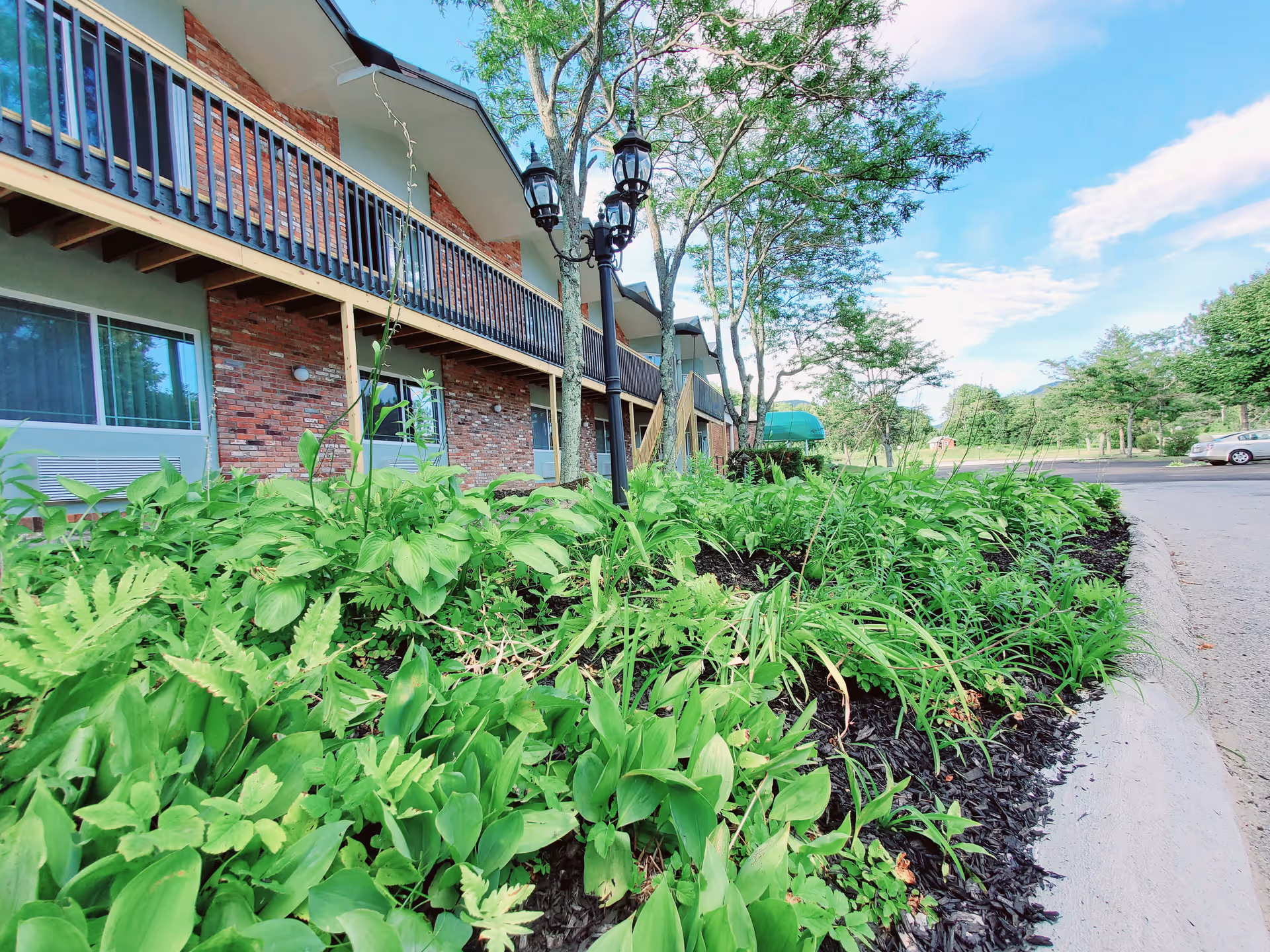 View of the exterior of a senior living facility building with a brick facade and balcony railing. In the foreground, there is a landscaped garden bed with various green plants and a black lamp post. Trees and a parking area with a car are visible in the background under a partly cloudy sky.
