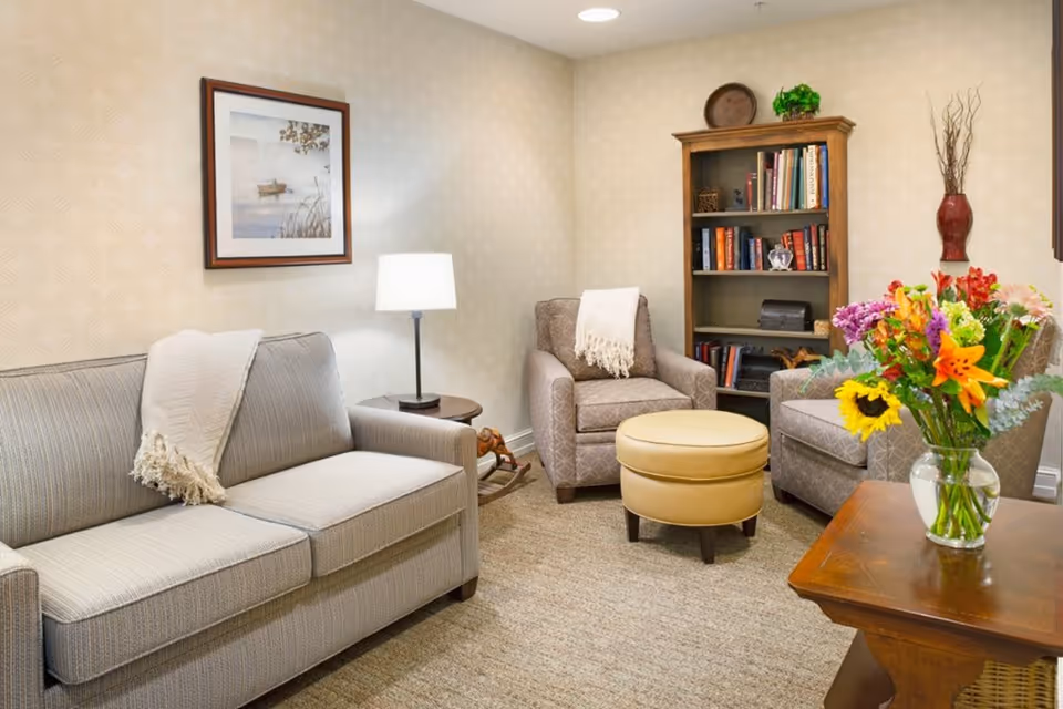 A cozy living room with a beige sofa and two patterned armchairs, each with a white throw blanket. A round beige ottoman is placed in the center. There is a wooden bookshelf filled with books and decorative items against the wall. A table lamp on a small side table illuminates the space. A framed picture hangs on the wall above the sofa, and a vase with a colorful bouquet of flowers sits on a wooden coffee table in the foreground.