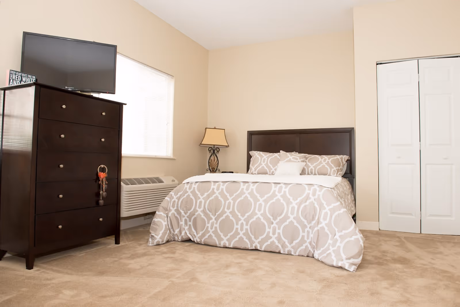 A bedroom with beige walls and carpet, featuring a bed with a patterned beige and white comforter and pillows, a dark wooden headboard, a matching dark wooden dresser with a flat-screen TV on top, a window with blinds, a decorative table lamp, and a white closet door.