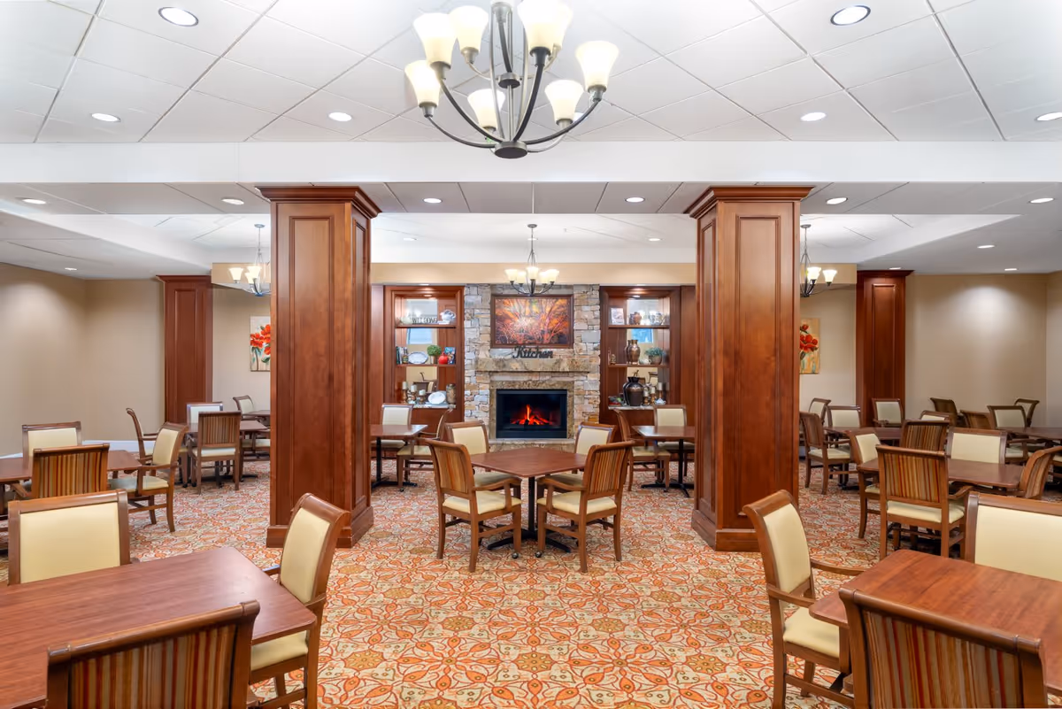 A spacious dining room with multiple wooden tables and chairs arranged neatly on a patterned carpet. The room features large wooden pillars, a stone fireplace with a fire burning, and built-in shelves displaying decorative items. Two chandeliers hang from the ceiling, providing warm lighting.