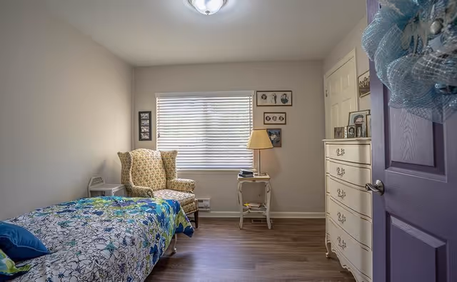 A cozy bedroom with a single bed covered in a floral bedspread, a patterned armchair near a window with blinds, a small side table with a lamp, and a white dresser. The walls are decorated with framed pictures, and the door is partially open with a decorative wreath hanging on it.