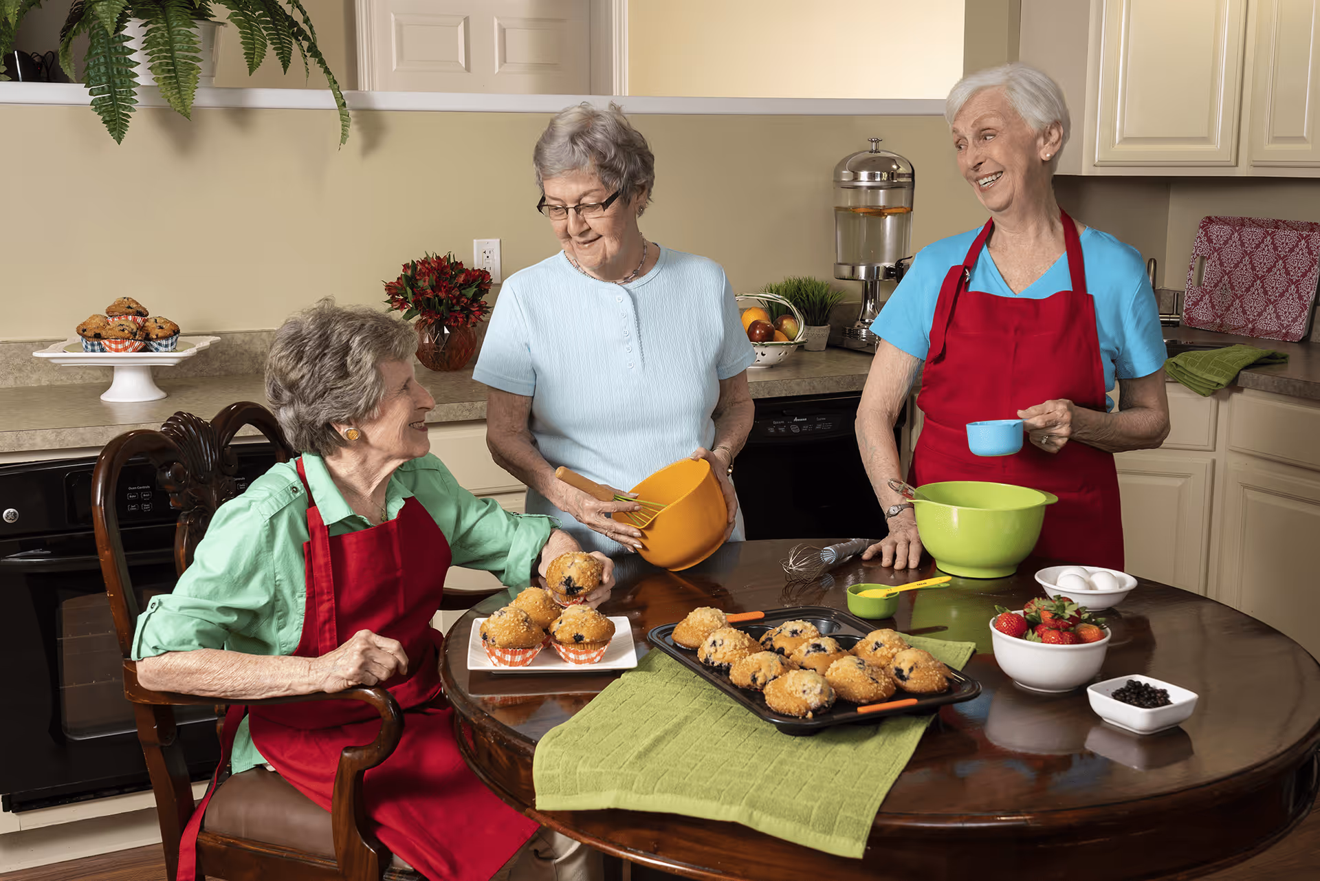 Three elderly women in a kitchen preparing and enjoying freshly baked muffins. Two women are wearing red aprons, one sitting and holding a muffin, the other standing and holding a blue measuring cup. The third woman, standing in the middle, is holding an orange mixing bowl. The kitchen counter and table have various baking items including a tray of muffins, bowls of strawberries, eggs, and blueberries.