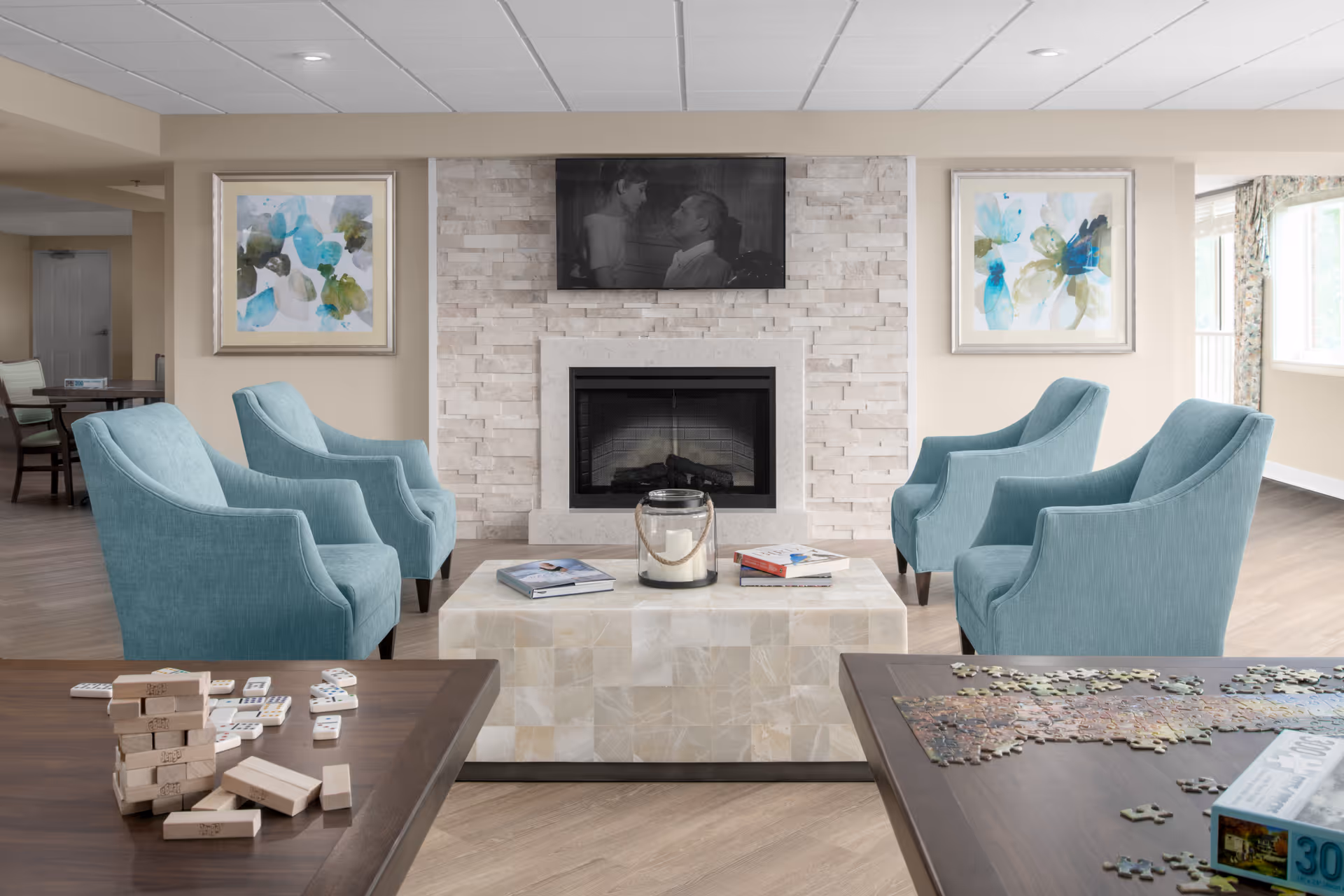 A cozy living room area with four light blue armchairs arranged around a square coffee table with a candle and books on it. Behind the table is a modern fireplace with a stone surround and a black and white photo displayed above it. Two abstract floral paintings hang on the walls on either side of the fireplace. In the foreground, there are two tables with a Jenga game and a partially completed jigsaw puzzle.