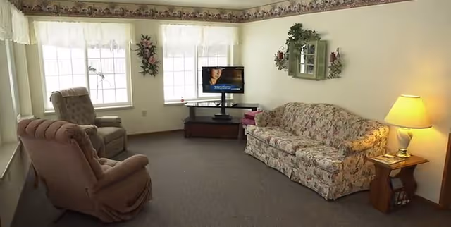 Small living room with a floral sofa, two recliners, a TV on a stand, lamp and large windows.
