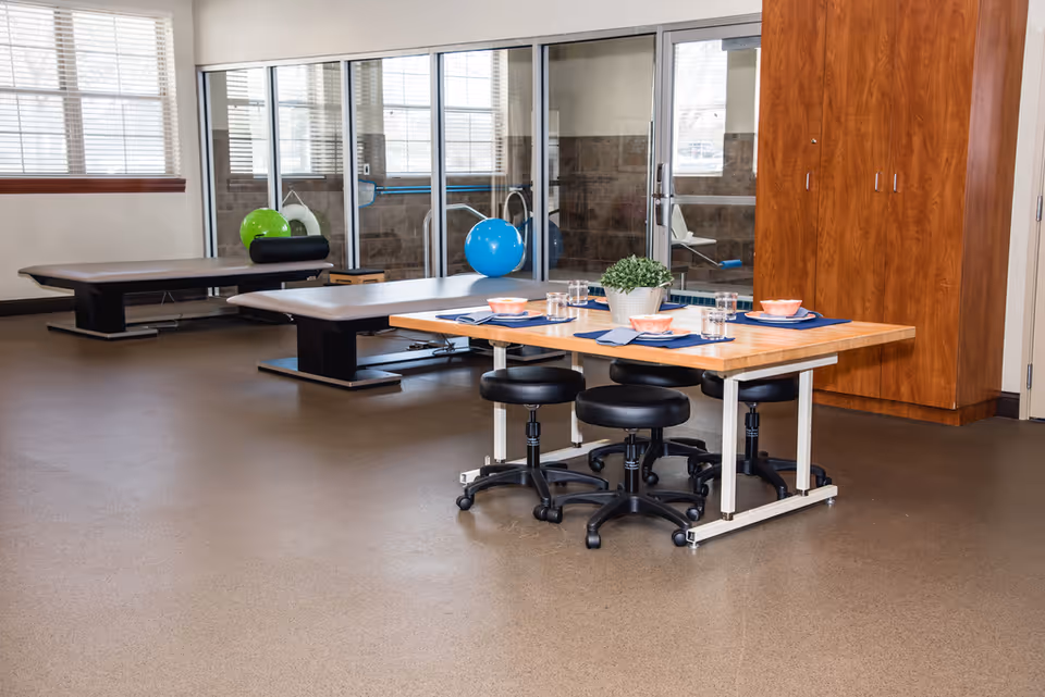 Multipurpose interior therapy room with a wooden table set with placemats and stools, treatment tables, exercise balls, and a glass partition.