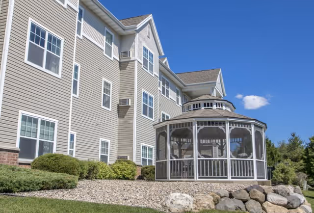 Exterior view of a multi-story senior living facility with beige siding and multiple windows. In the foreground, there is a white screened gazebo surrounded by rocks and landscaping under a clear blue sky.