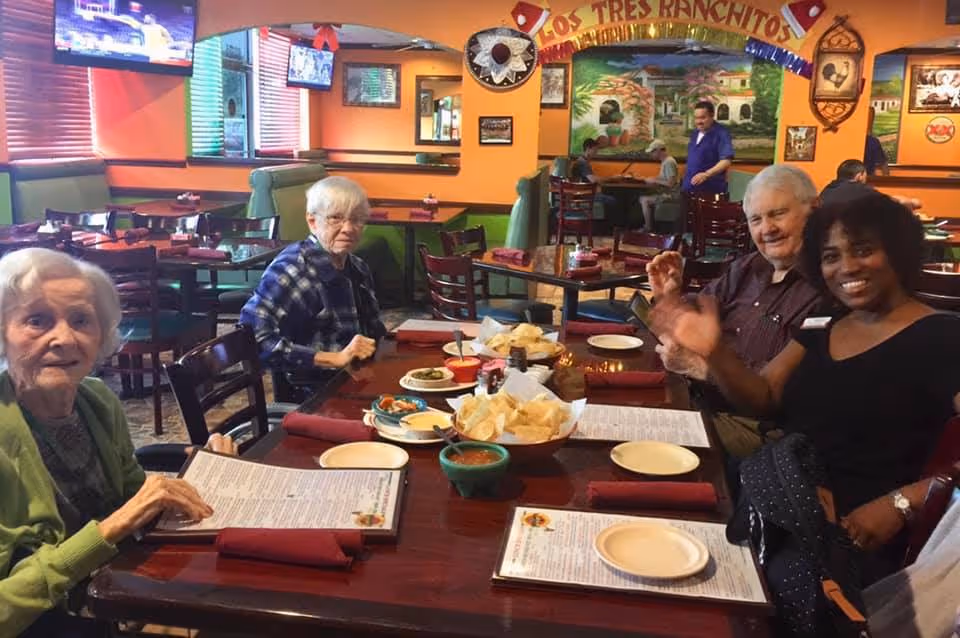 Four people sit at a table in a colorful restaurant dining room with menus, bowls of chips and salsa, and TV screens on the walls.