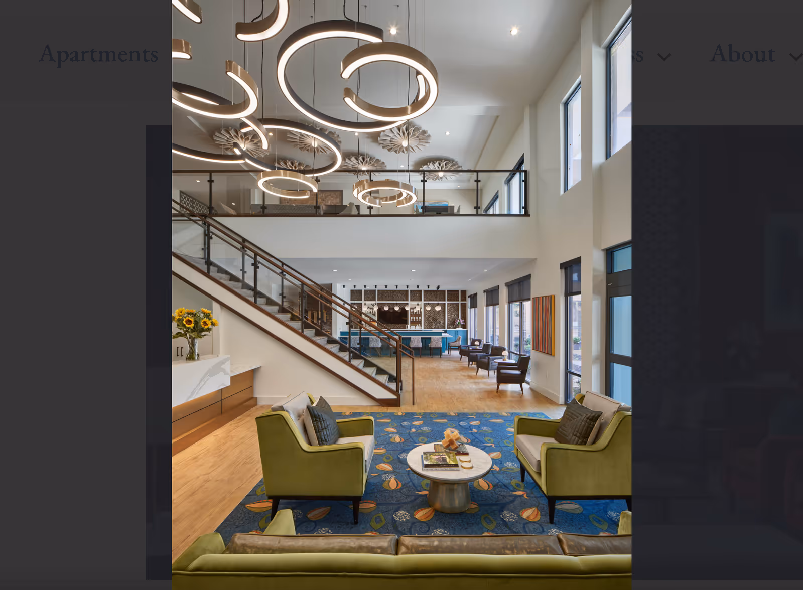 A spacious and modern living room area with high ceilings and large windows allowing natural light. The room features green armchairs and a sofa arranged around a small round coffee table on a blue patterned rug. A staircase with glass and wood railing leads to an upper level with additional seating. Contemporary circular pendant lights hang from the ceiling, and there are sunflowers in a vase on a side counter.