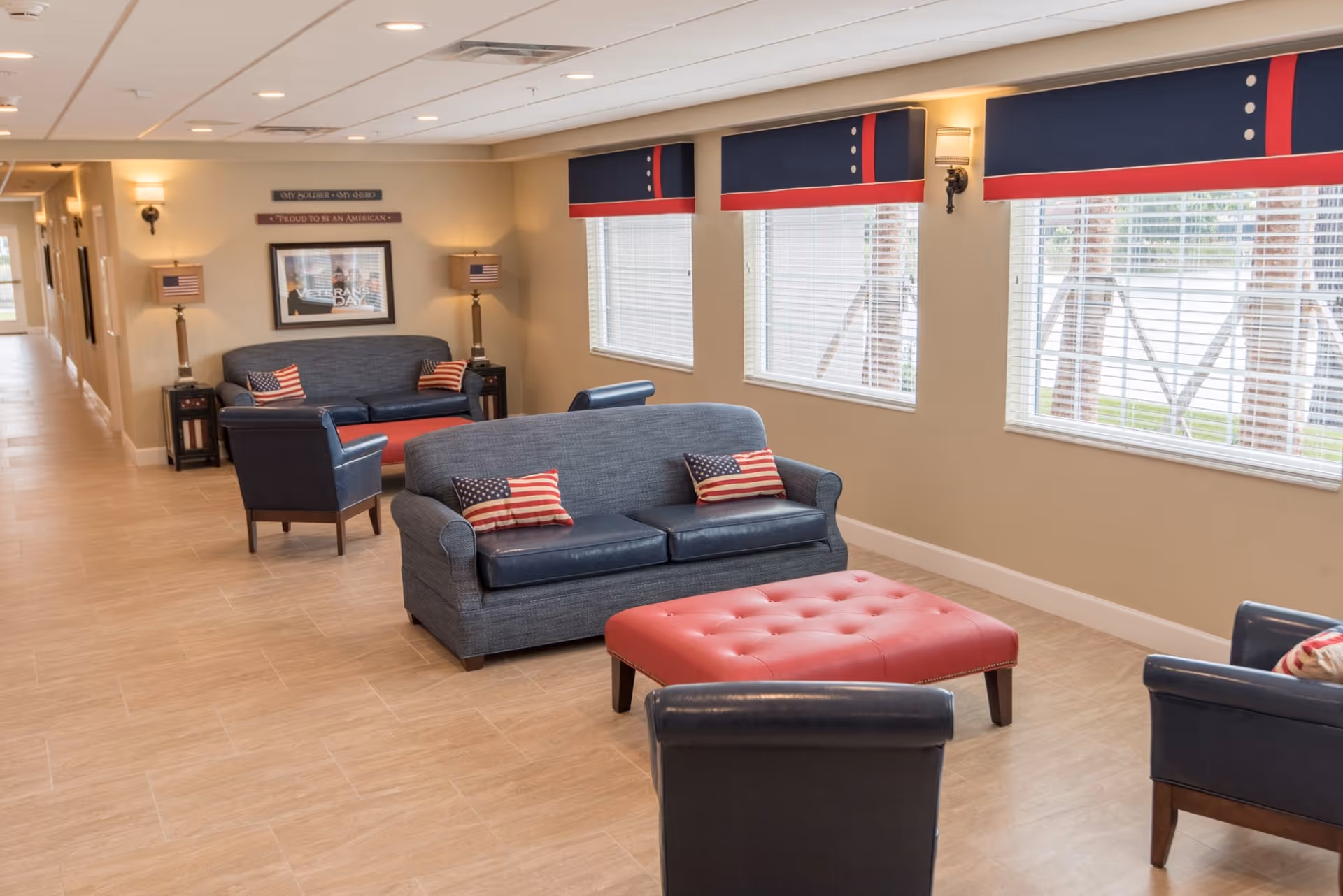 Bright communal lounge with sofas, armchairs, a red ottoman, American-flag pillows, and large windows.
