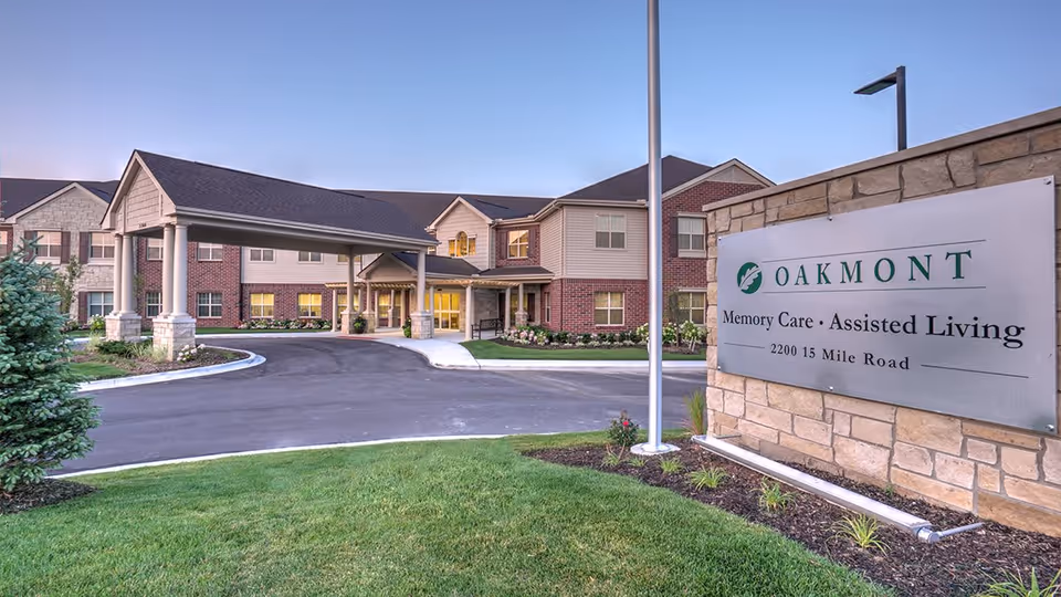 Exterior view of a senior living facility building with a covered entrance driveway, well-maintained landscaping, and a large sign that reads 'OAKMONT Memory Care • Assisted Living 2200 15 Mile Road'. The sky is clear and the building has a combination of brick and siding.