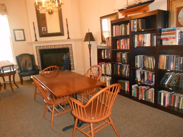 A cozy interior room featuring a wooden table with six matching wooden chairs around it. To the right, there are tall bookshelves filled with books. In the background, there is a fireplace with a mirror above it, flanked by two candlesticks. A floor lamp stands next to the bookshelves, and a chandelier hangs from the ceiling. The room has a warm and inviting atmosphere.