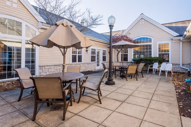 Patio courtyard with round tables, umbrellas, chairs, and a lamppost beside the exterior of a beige senior living building.