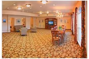 Spacious common area in a senior living facility with patterned carpet, several armchairs arranged near the walls, a wooden dining table with chairs, a chandelier, and large windows with curtains allowing natural light.