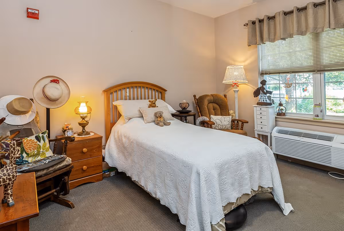 A cozy bedroom with a single bed covered in a white quilt, two teddy bears on the bed, a wooden headboard, a wooden nightstand with a vintage lamp, a rocking chair with cushions, a window with blinds and curtains, and various decorative items including hats and figurines.