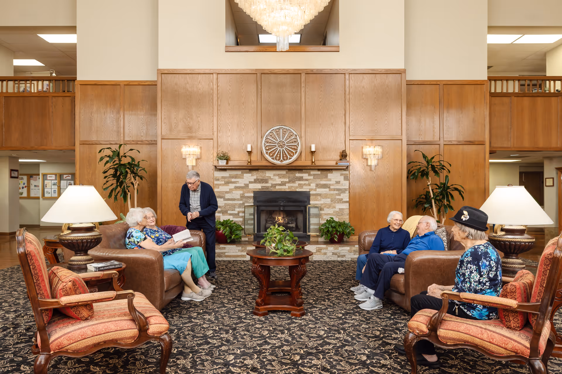 A cozy common area in a senior living facility with six elderly people sitting and chatting around a fireplace. The room features wooden paneling, a patterned carpet, two leather sofas, two upholstered armchairs, table lamps, and potted plants. A large chandelier hangs from the ceiling.